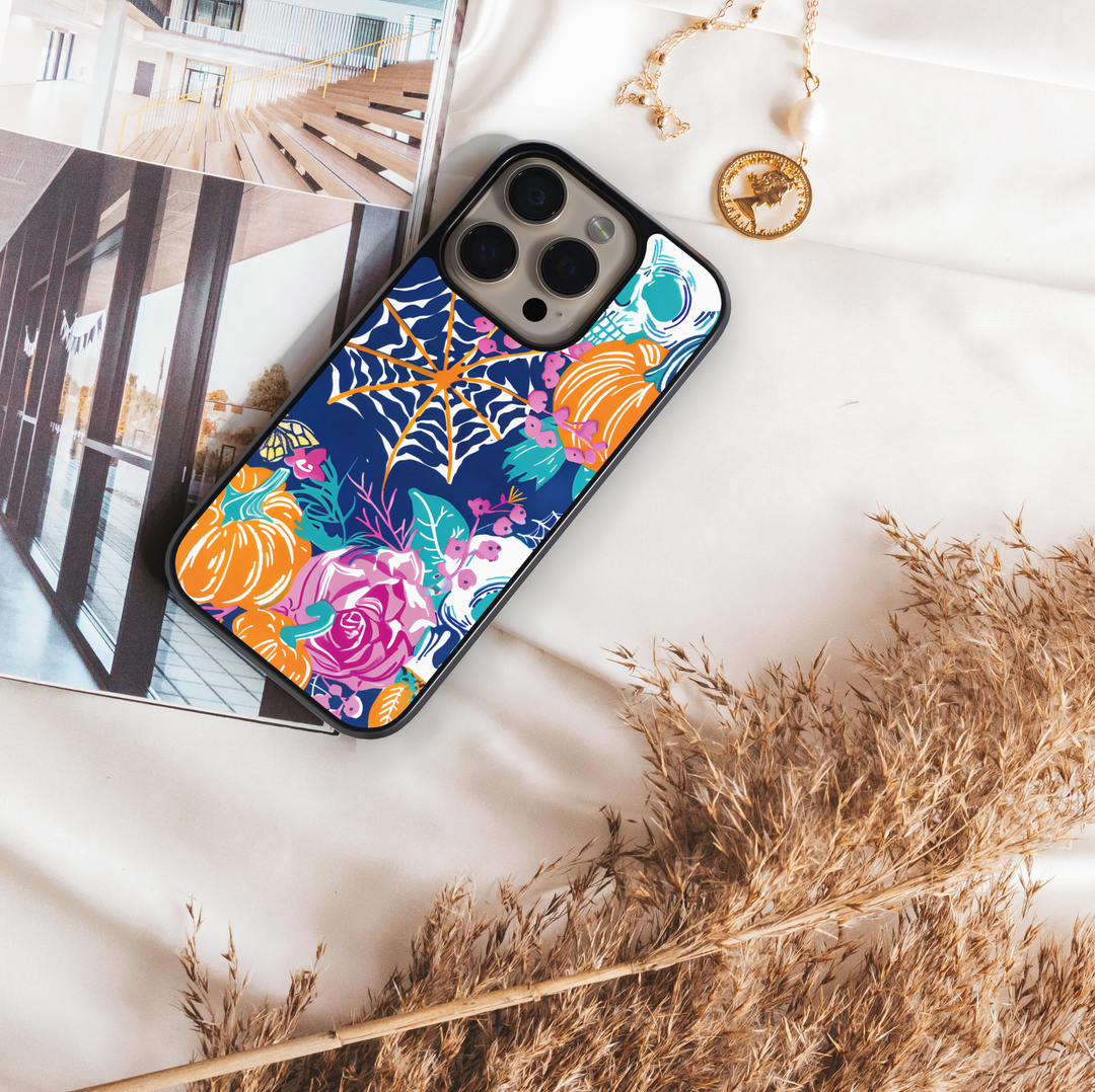 Midnight Bloom iPhone case with pumpkins, skull, spider web, and colorful floral design on navy background.