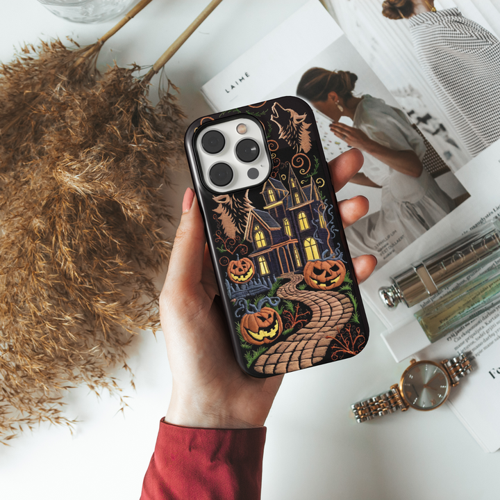 Embroidered haunted house iPhone case with glowing jack-o'-lanterns, spooky trees, howling wolves, and a winding path on a black background. Halloween-ready.