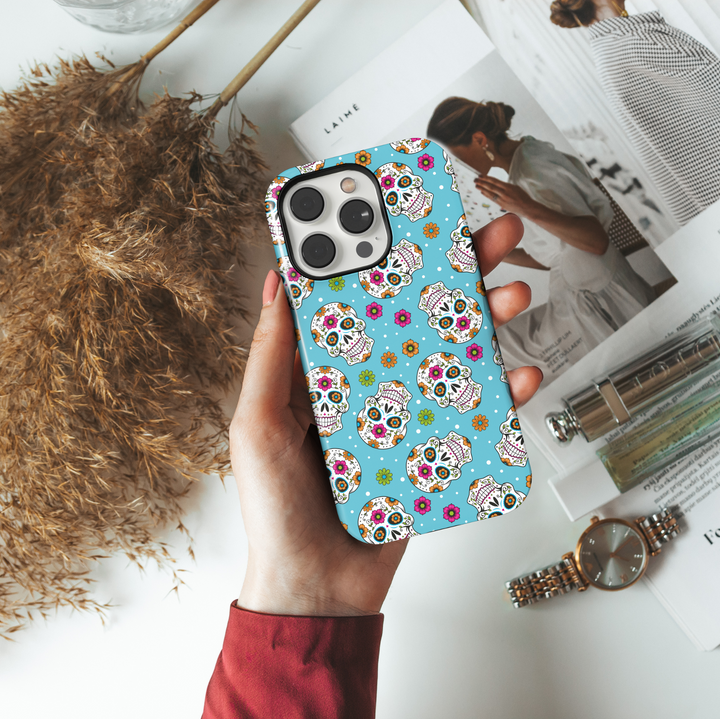 Bright sugar skull iPhone case with vibrant orange, pink, green, and blue floral patterns on a turquoise blue background. Features traditional Day of the Dead (Día de los Muertos) skull art for iPhone 16 Pro Max, 15, 14, and 13 models. Bold, festive, and eye-catching design perfect for Halloween and fall celebrations.

