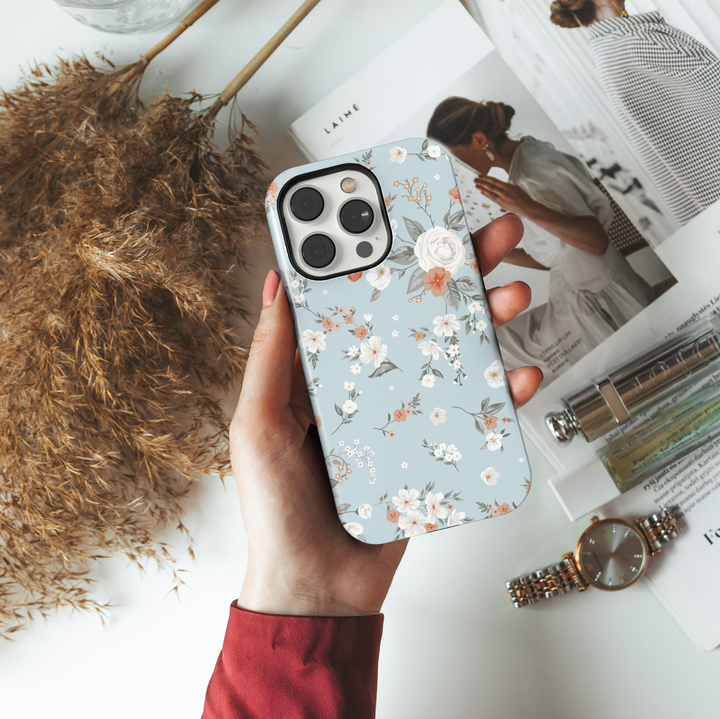 iPhone case with an elegant vintage floral design featuring ivory roses, orange blossoms, and soft greenery on a muted dusty blue background.