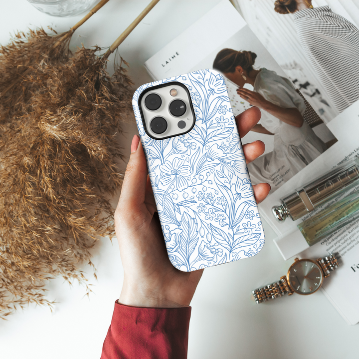White iPhone case featuring a detailed blue floral line art pattern with various wildflowers, leaves, and berries, creating a clean and elegant botanical look.