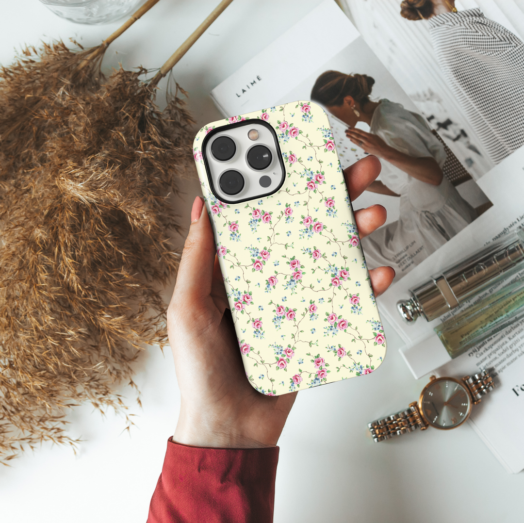 Cream iPhone case adorned with a vintage-inspired pattern of delicate pink roses and tiny blue florals connected by thin trailing vines, creating a romantic, timeless aesthetic.