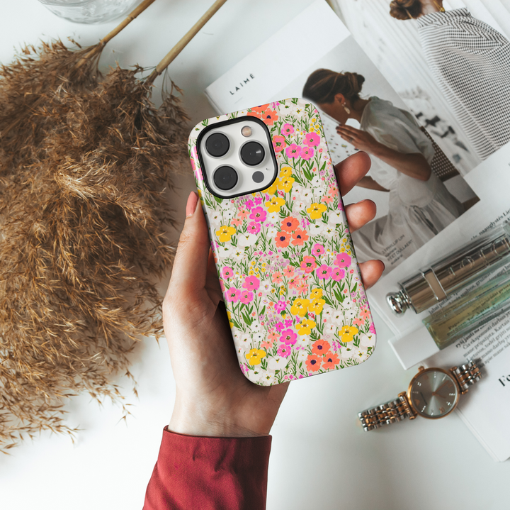 Charming iPhone case with a vibrant wildflower meadow pattern featuring hand-painted flowers in pink, yellow, coral, and white on a soft cream background.
