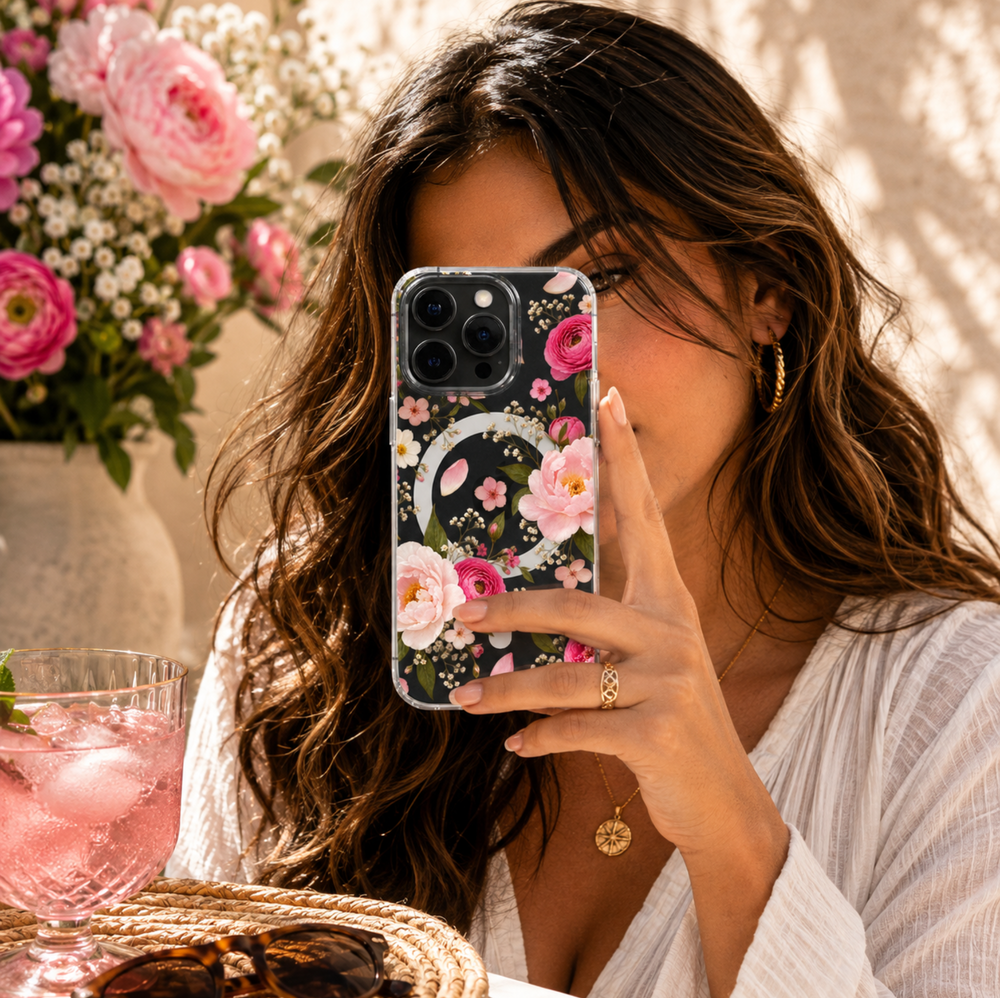 Clear MagSafe iPhone case featuring blush pink and hot pink peonies, delicate white flowers, green leaves and scattered petals.