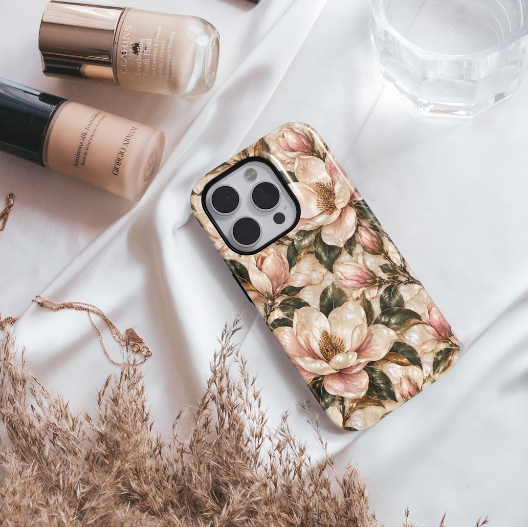 January birth flower iPhone 18 Pro Max case featuring Magnolia Grace with elegant ivory and blush pink magnolia blossoms, golden botanical accents, and deep green leaves on a champagne-inspired background with a glossy printed finish.