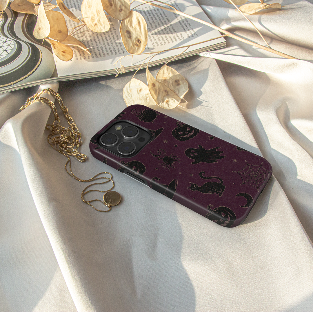 Midnight Witchery iPhone case with black cats, ghosts, pumpkins, witch hats, cauldrons, spiders and moons on a dark plum Halloween background.