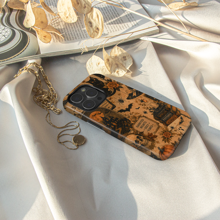 A phone case with a vintage Halloween-themed print featuring tombstones, pumpkins, bats, and spiderwebs, styled on draped white fabric with a gold chain necklace, dried leaves, and an open book.