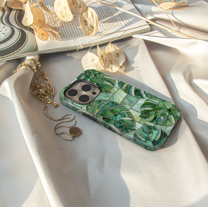 Green stained glass iPhone case featuring layered monstera leaves in emerald, jade, and sage botanical mosaic artwork.