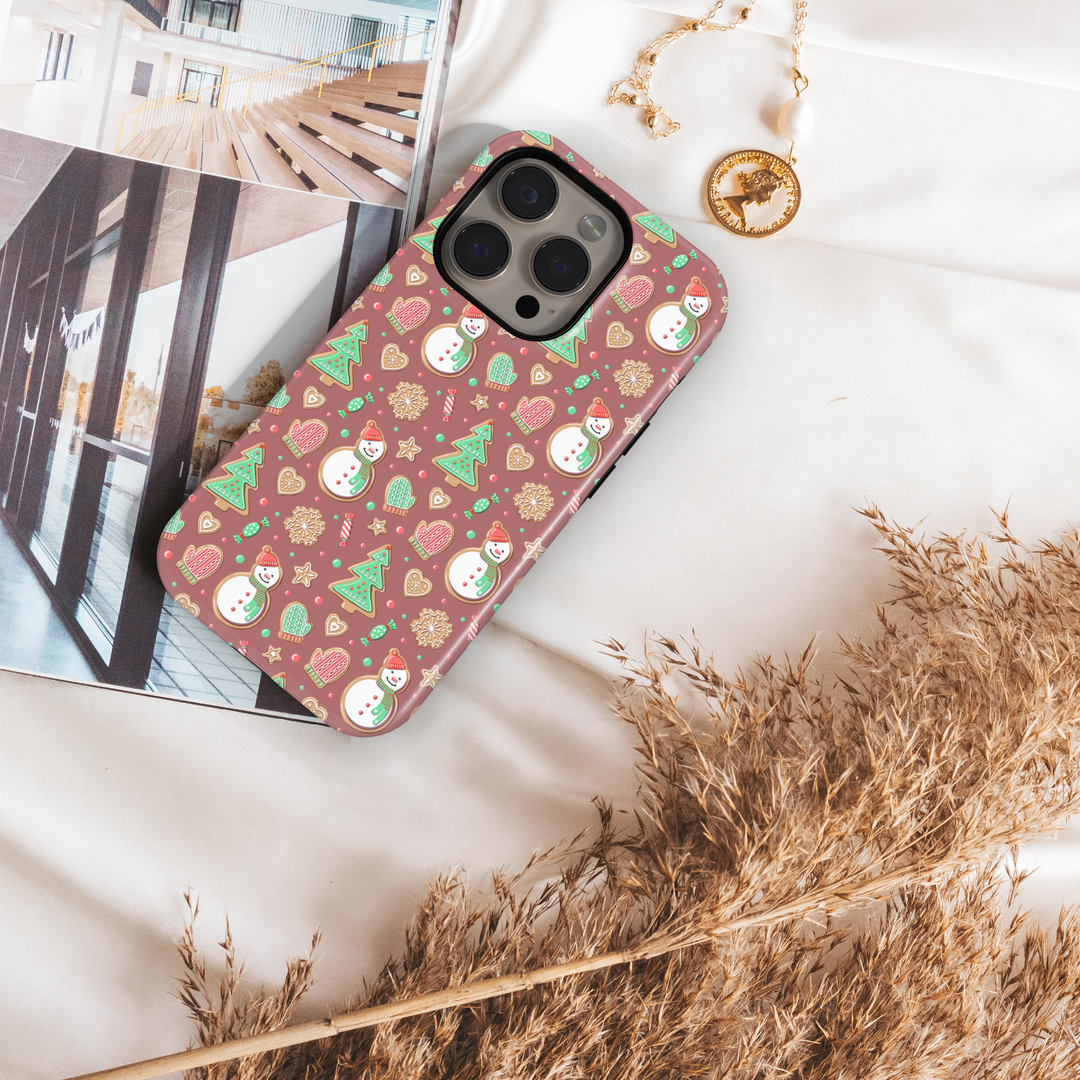 Cute Christmas iPhone case with gingerbread snowmen, holiday cookies, mittens, and festive icons on a warm pink background